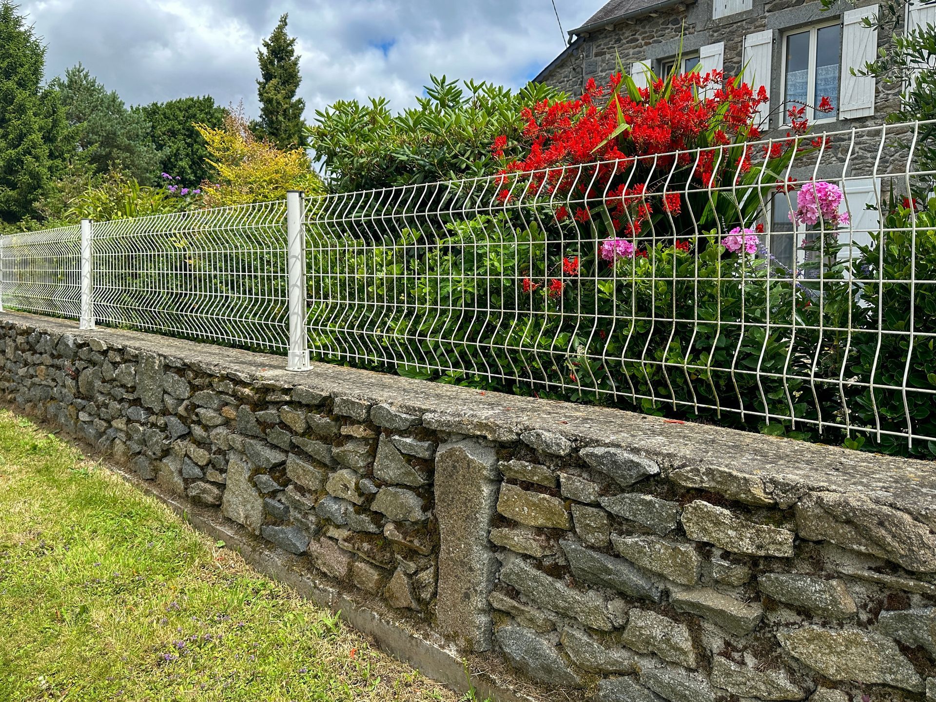 Stone wall topped with a white wire fence. Behind it, a house with pink and red flowers.