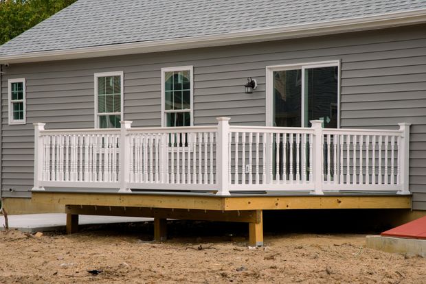 White-railed deck attached to a gray house with windows and sliding glass door. Deck is over a wooden frame, in yard.