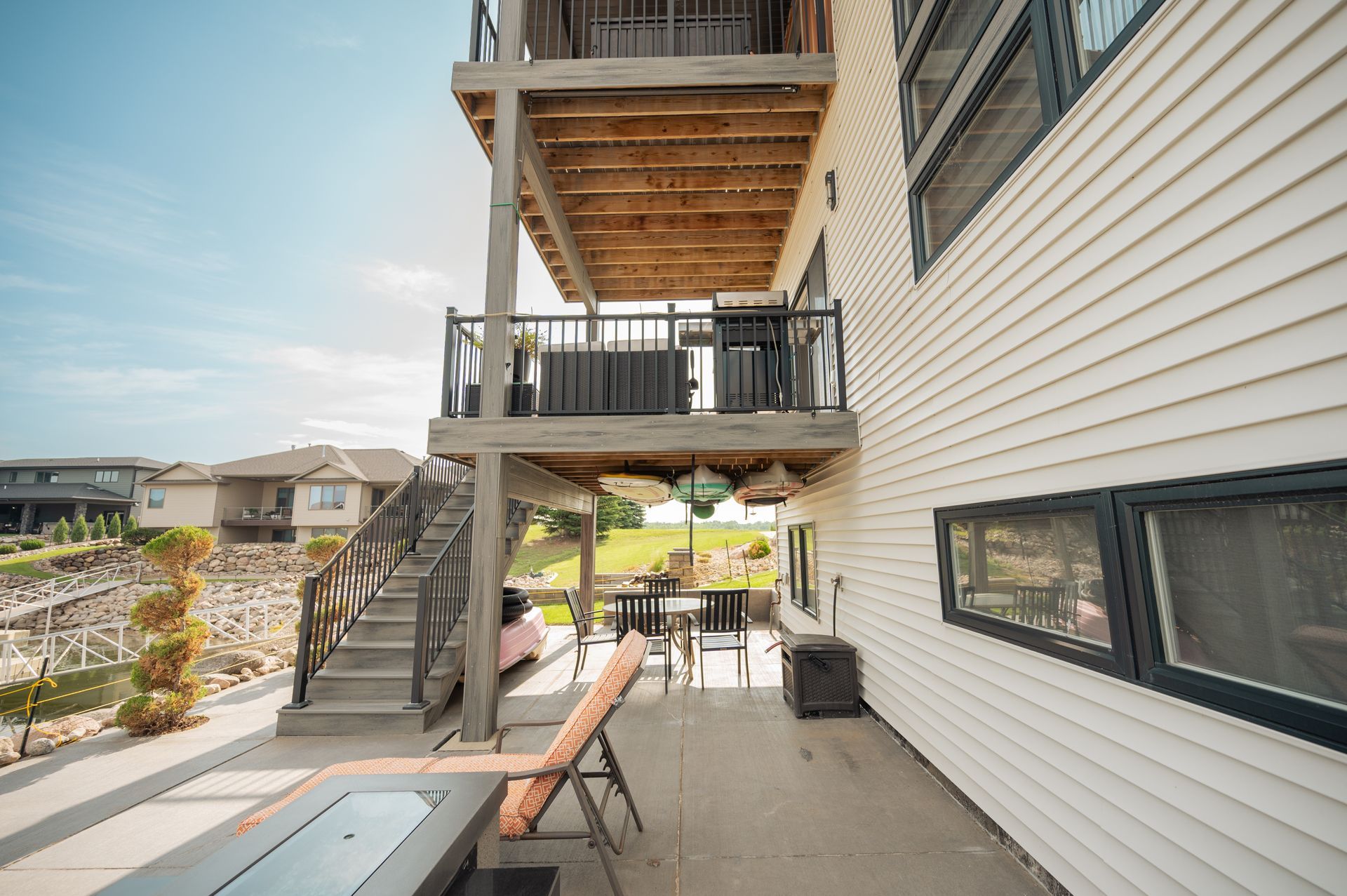 Two-story house with decks overlooking a lake. Gray stairs lead up. Patio with furniture and fire pit.