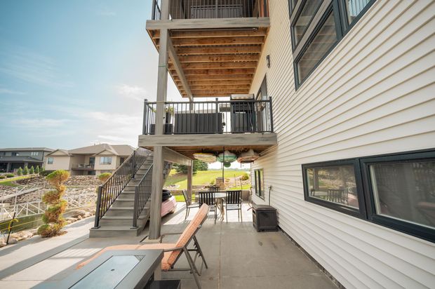 Two-story house with decks overlooking a lake. Gray stairs lead up. Patio with furniture and fire pit.