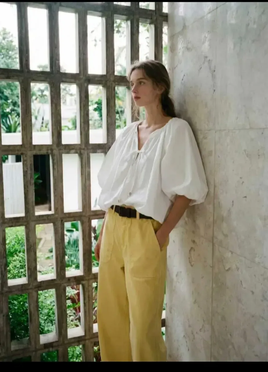 Une personne vêtue d'un chemisier blanc et d'un pantalon jaune, appuyée contre une paroi vitrée.