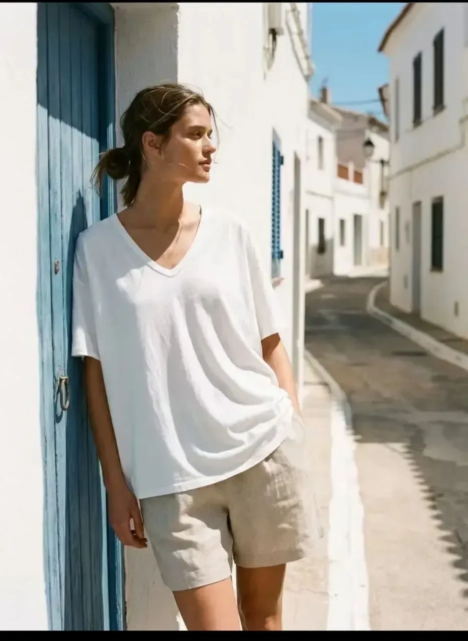 Une femme en haut blanc et short beige se tient près d'une porte bleue dans une rue étroite et ensoleillée.