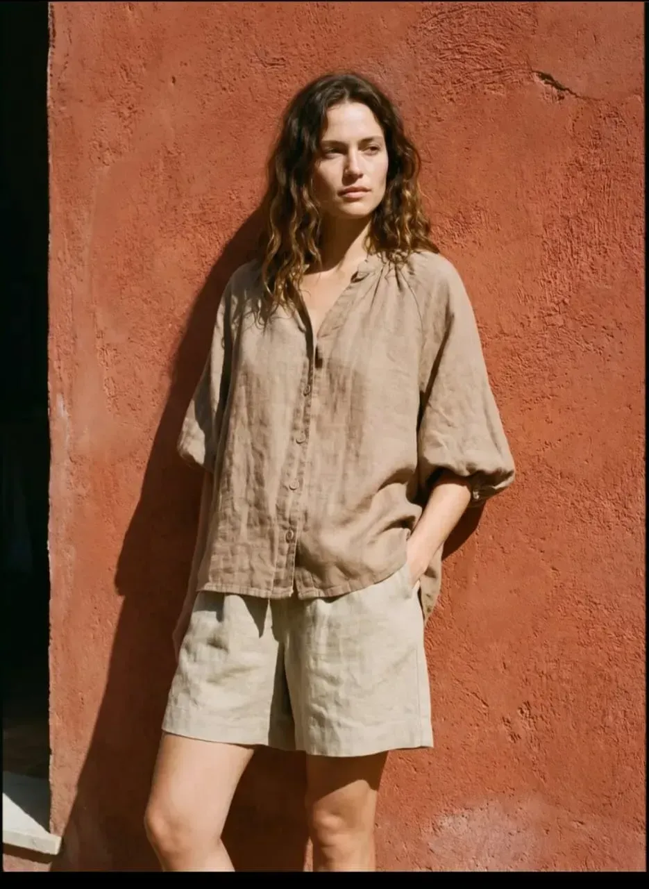 Une personne en chemise beige et short est appuyée contre un mur rouge au soleil.