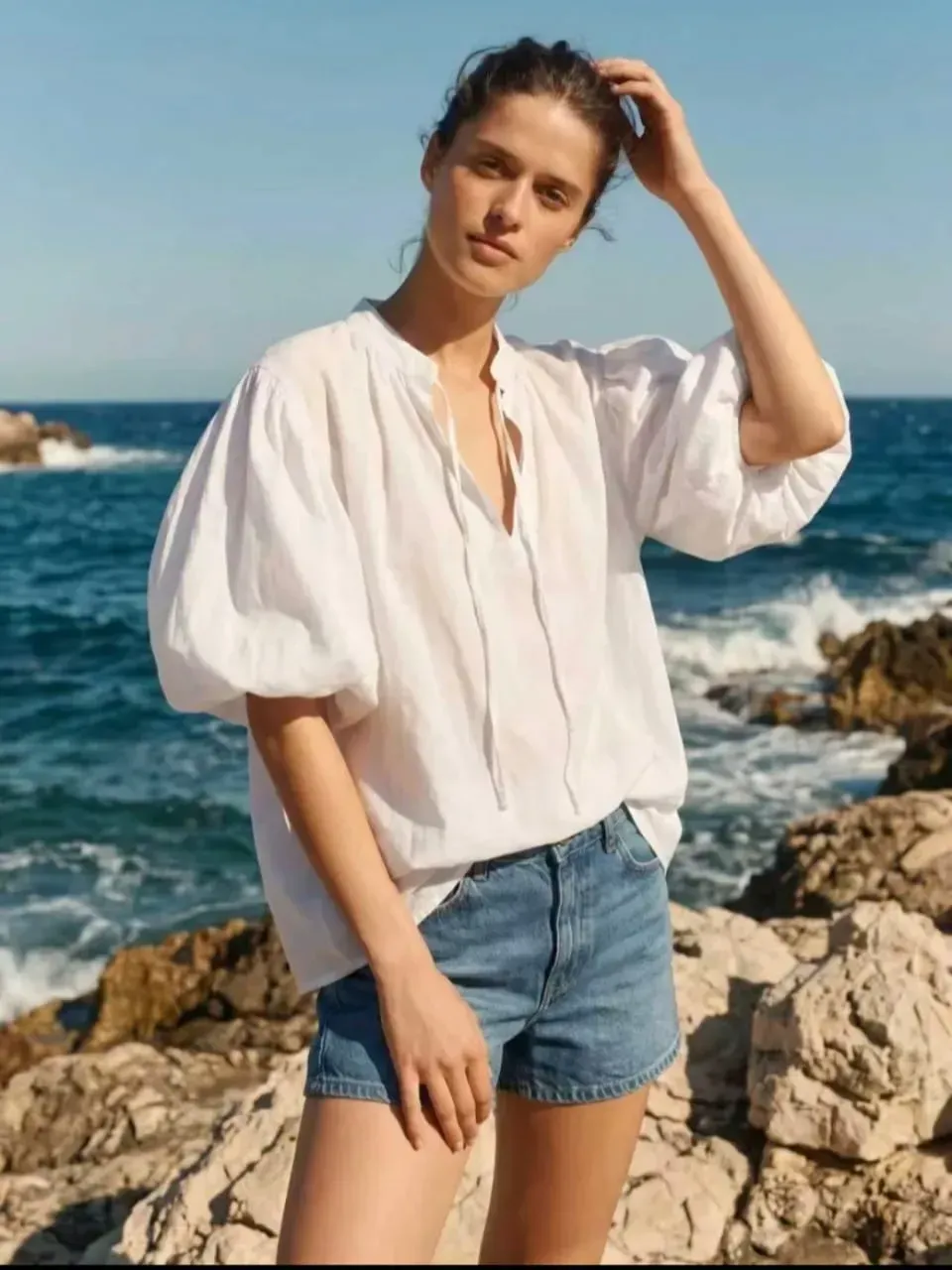 Une femme en chemisier blanc et short en jean se tient debout sur un rivage rocheux au bord de l'océan.