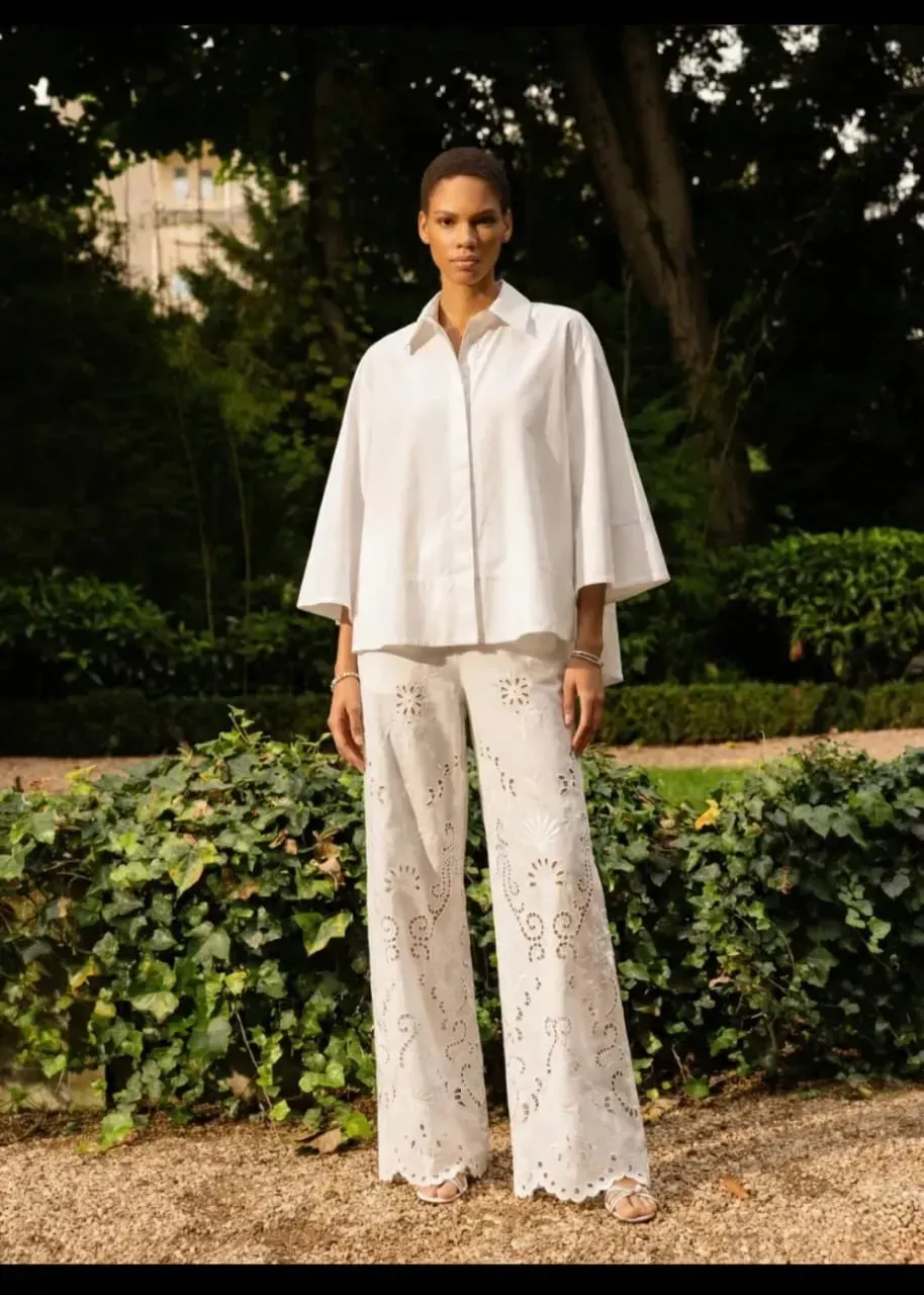 Une personne se tient debout à l'extérieur, vêtue d'une chemise blanche et d'un pantalon à motifs, dans un jardin.