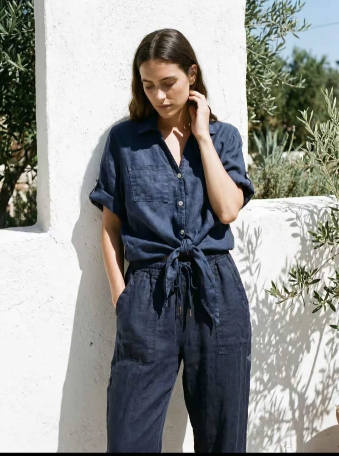 Personne en combinaison pantalon bleu marine ceinturée, debout près d'un mur blanc à l'extérieur, regardant vers le bas.