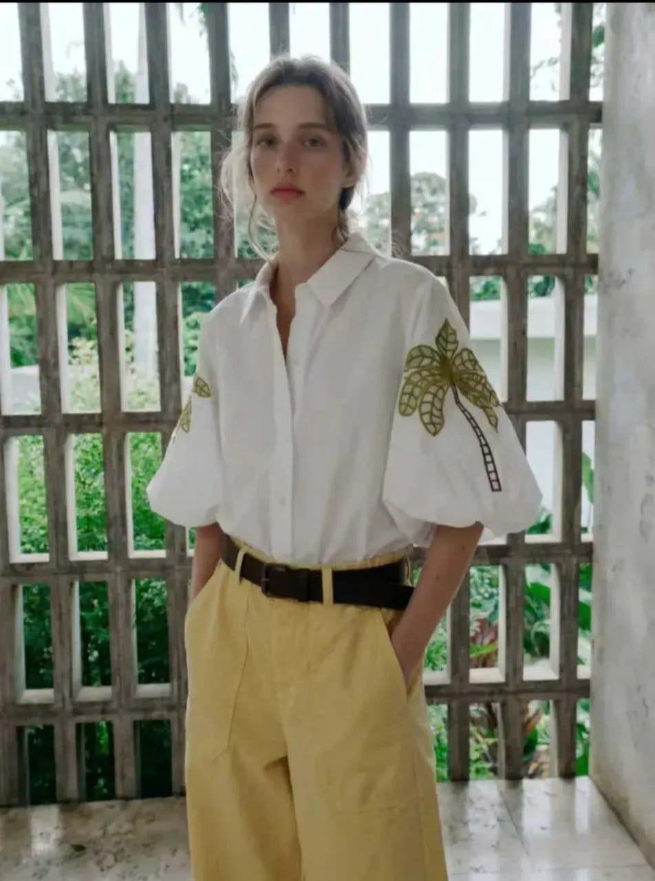 Une personne en chemise blanche et pantalon beige se tient près d'une fenêtre à barreaux dans une pièce lumineuse.