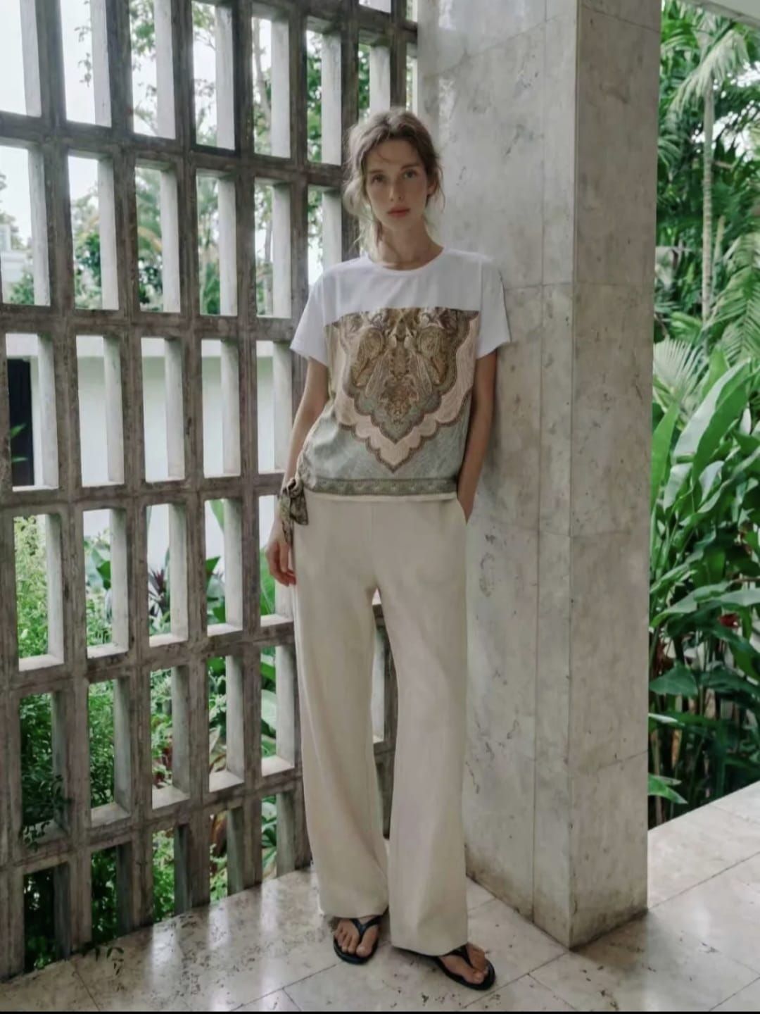 Une personne se tient près d'un pilier blanc sur une terrasse carrelée, portant un t-shirt blanc à motifs