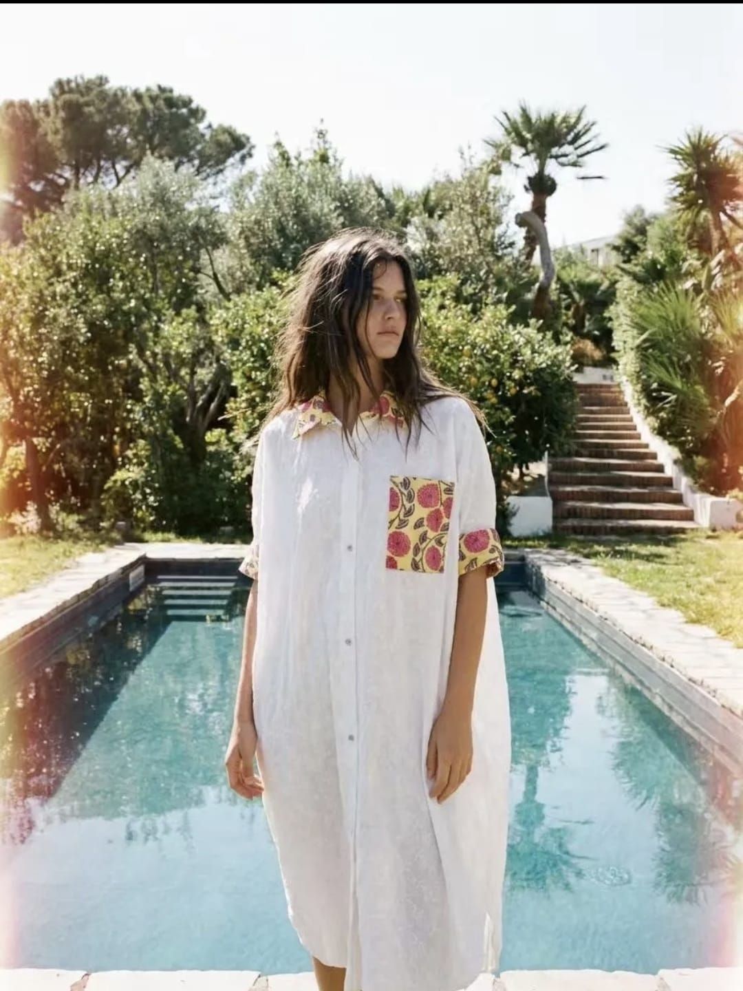 Femme en robe blanche debout au bord d'une piscine turquoise avec une végétation luxuriante et des marches en arrière-plan