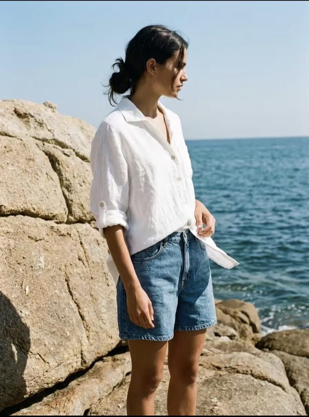 Une personne se tient près d'un mur vitré, vêtue d'un t-shirt blanc, d'un pantalon beige et de sandales