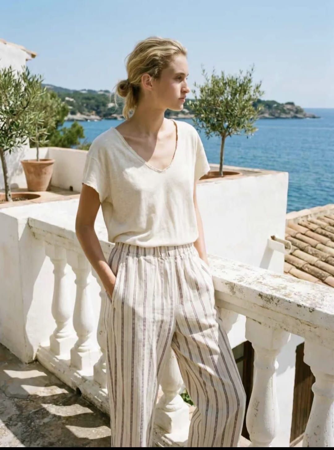 Femme en chemisier blanc et pantalon rayé sur une terrasse en bord de mer, avec eau bleue et arbres
