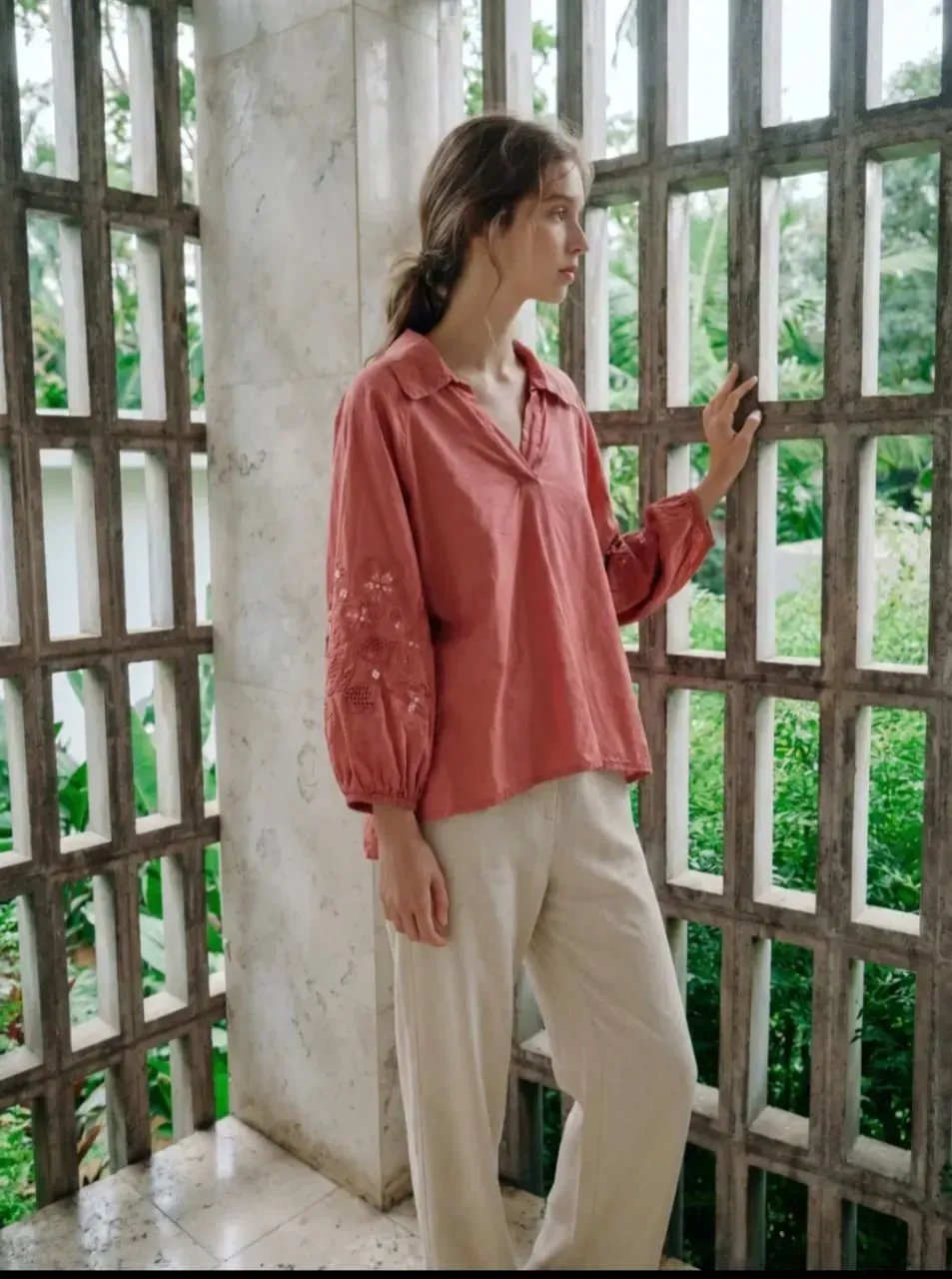 Une personne vêtue d'une chemise rose et d'un pantalon clair se tient près d'une grande fenêtre et regarde dehors.
