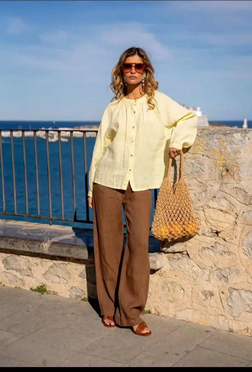 Personne debout près d'une rambarde en bord de mer, vêtue d'une chemise jaune pâle et d'un pantalon marron.