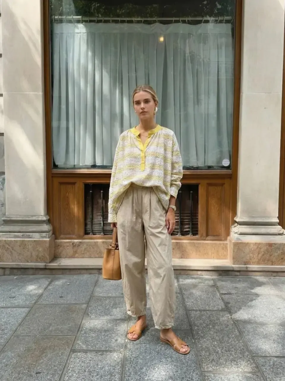 Une personne debout sur le trottoir, vêtue d'un pantalon beige et d'un chemisier jaune à motifs, devant une vitrine.