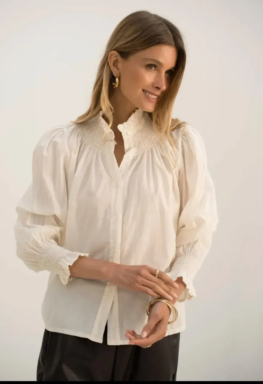 Femme en chemisier blanc et pantalon noir posant sur fond clair, tenant des lunettes.