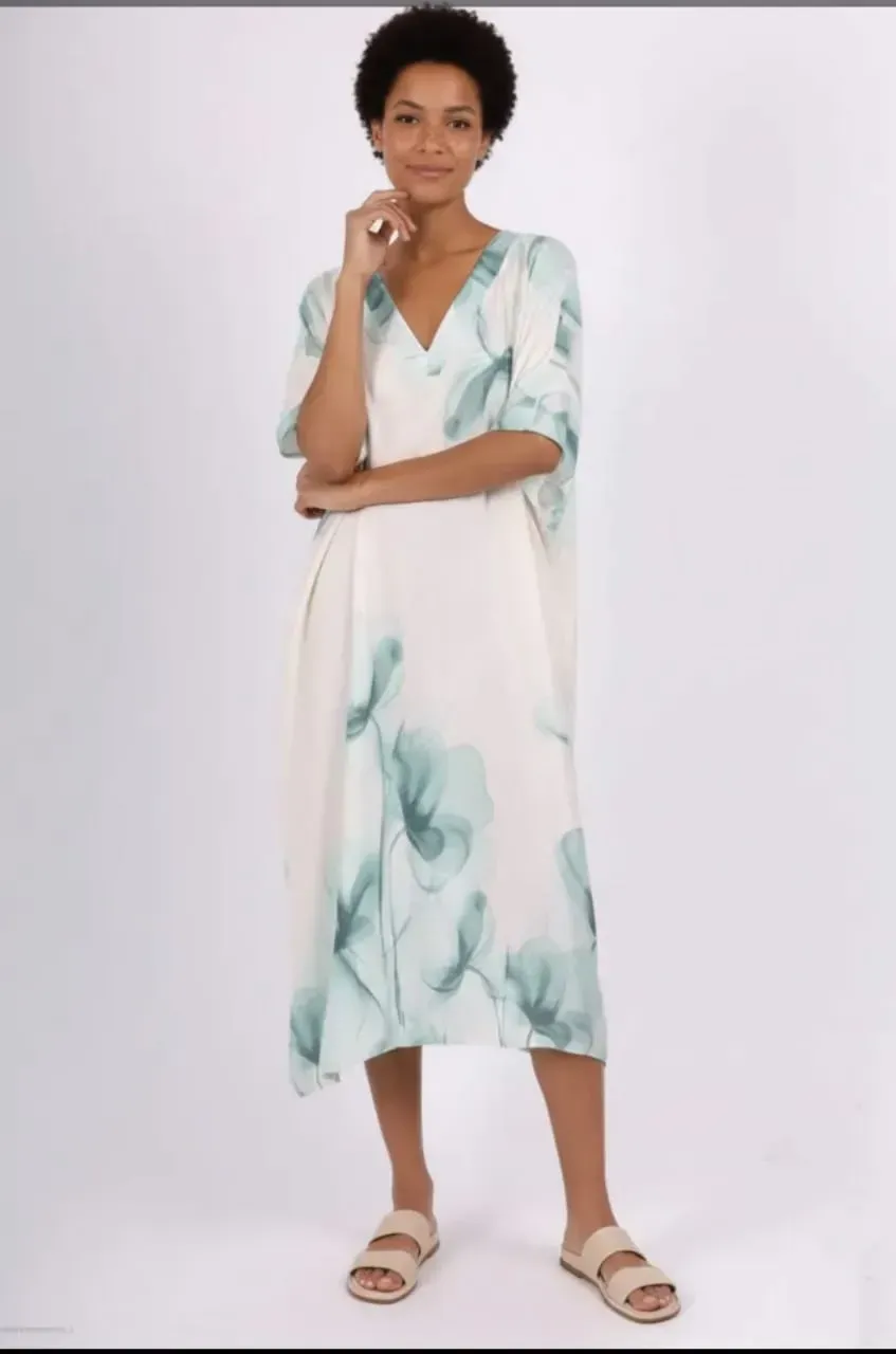 Femme en robe fleurie blanche et menthe et sandales, se tenant sur un fond uni.