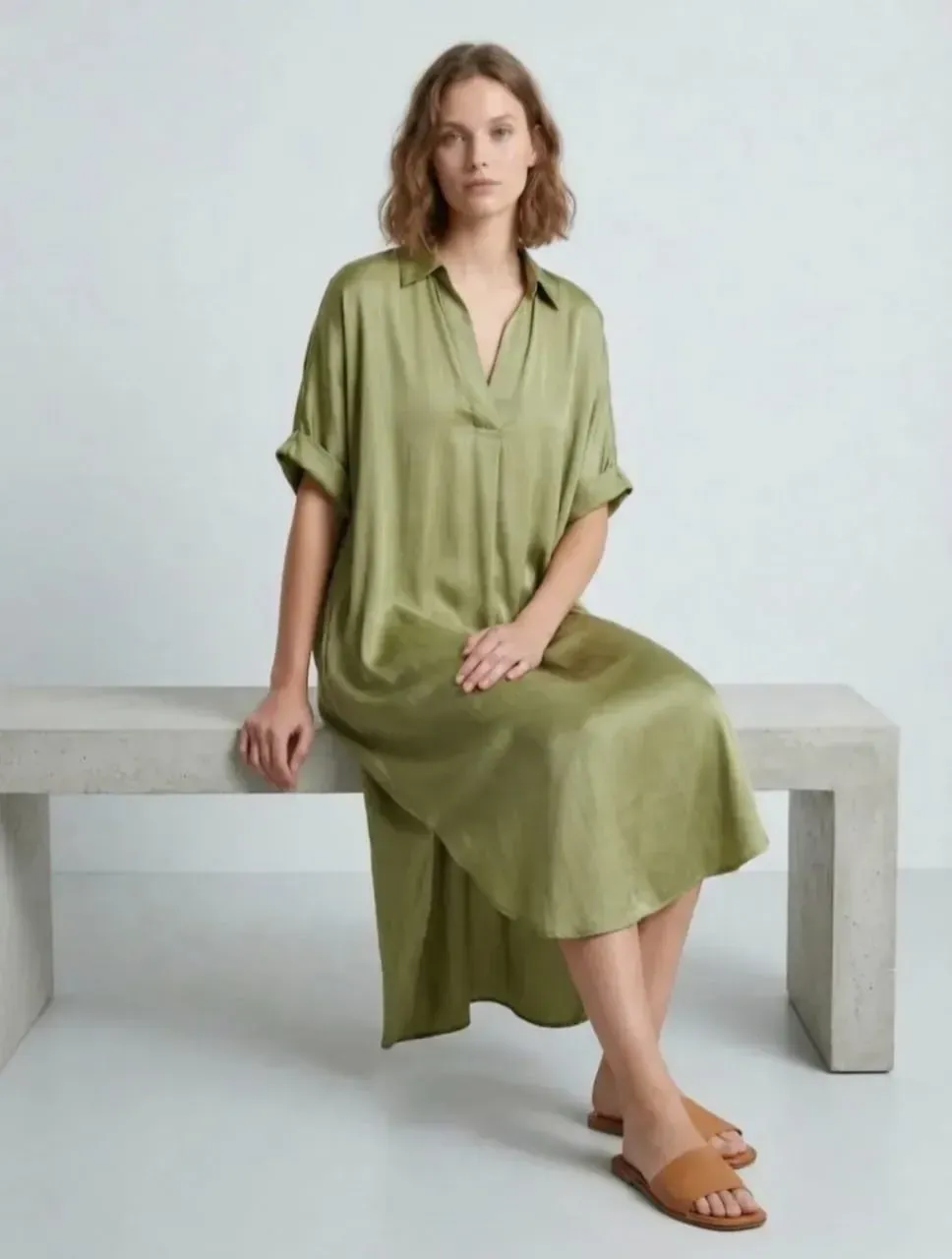Femme en robe vert olive assise sur un banc en béton sur fond gris clair.