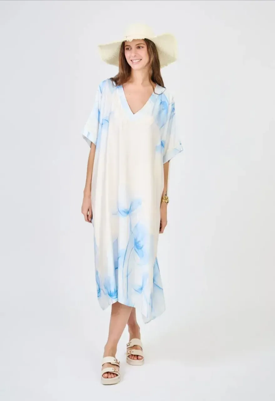Femme portant une robe longue bleu clair et blanche, un chapeau de paille et des sandales, se détachant sur un fond blanc.