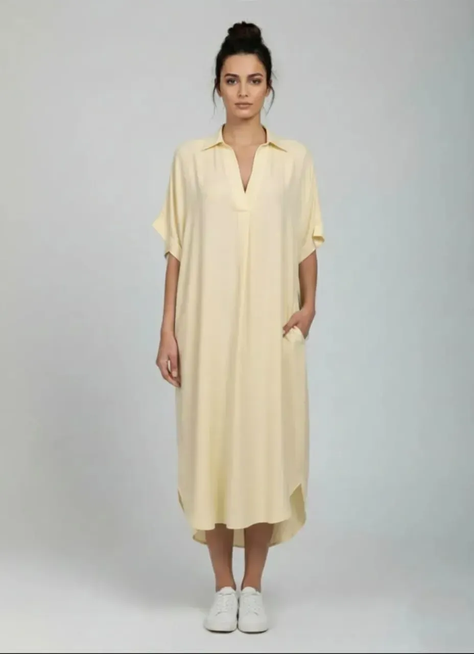 Femme debout, vêtue d'une robe ample jaune pâle et de baskets blanches, sur fond gris.