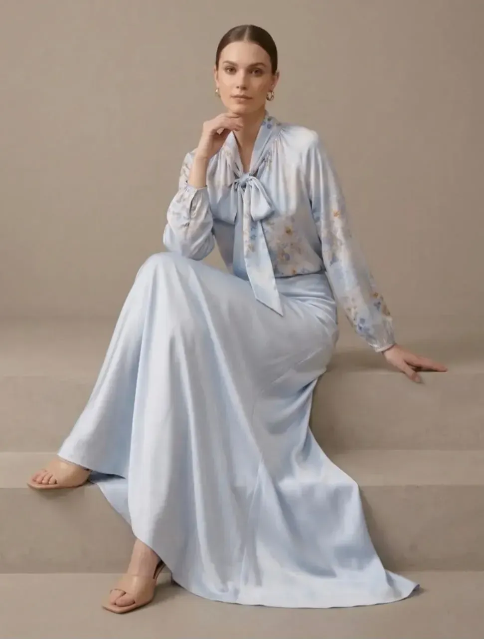 Femme en robe bleu clair assise sur une marche beige devant un fond neutre.