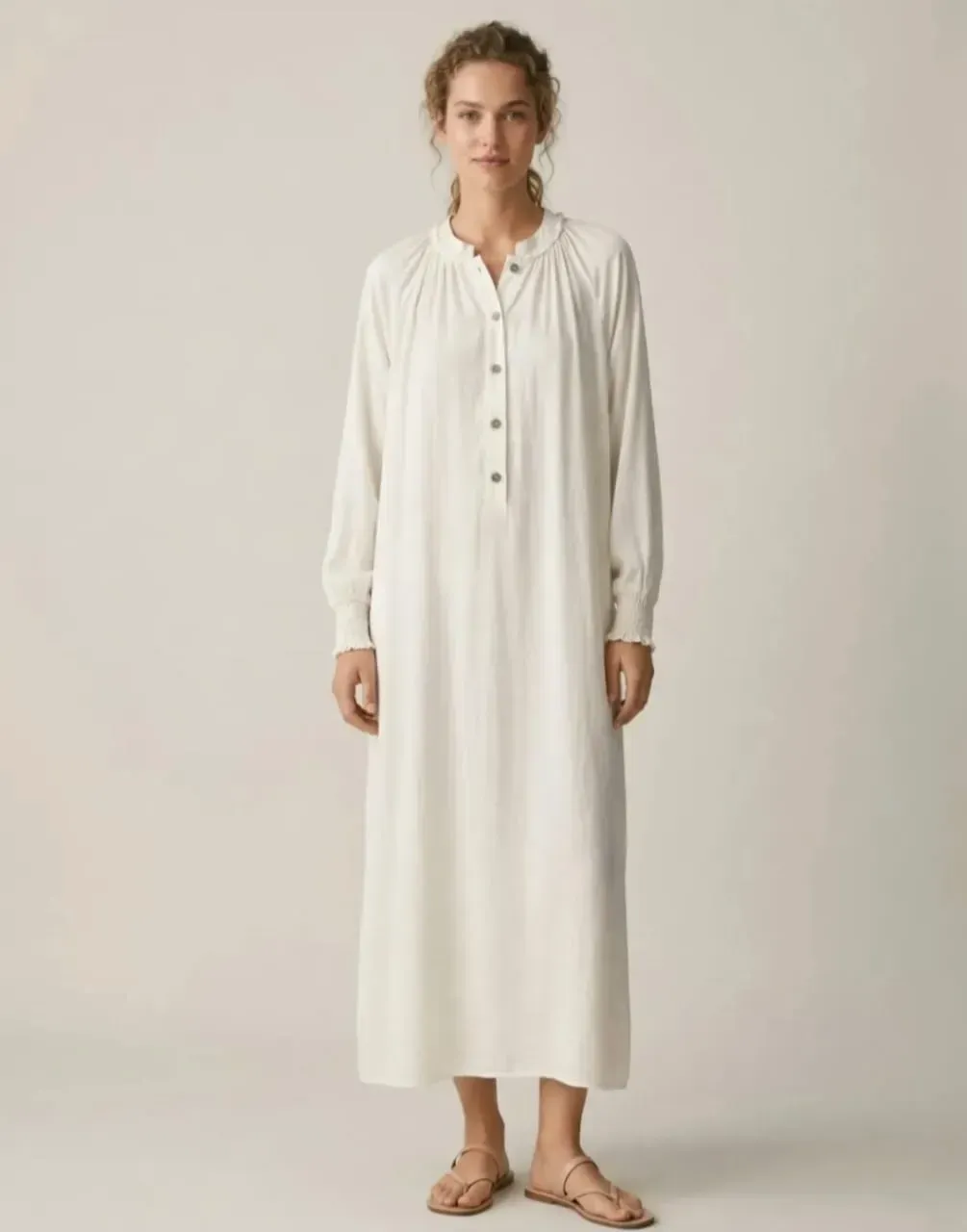 Personne vêtue d'une longue robe blanche boutonnée sur le devant, debout pieds nus sur un fond uni