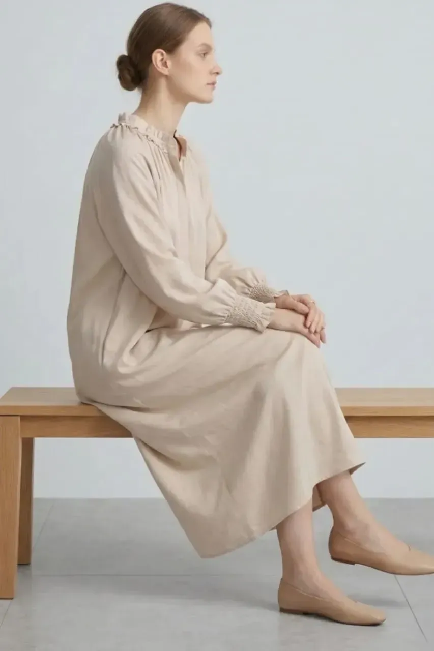 Une personne vêtue d'une robe beige est assise de profil sur un banc en bois, sur un fond gris clair.