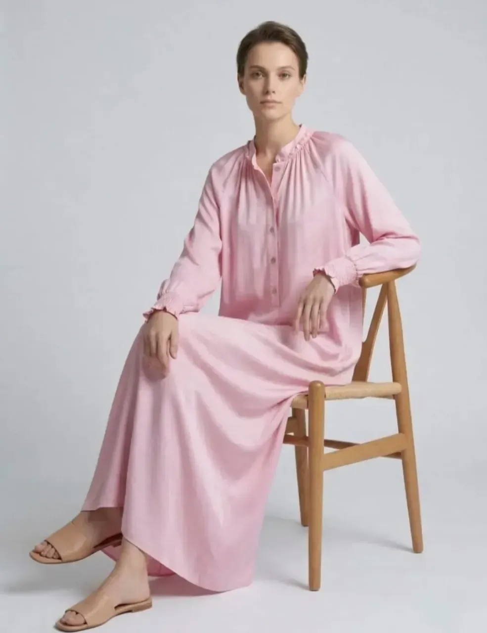 Femme en robe rose assise sur une chaise en bois devant un fond gris clair.