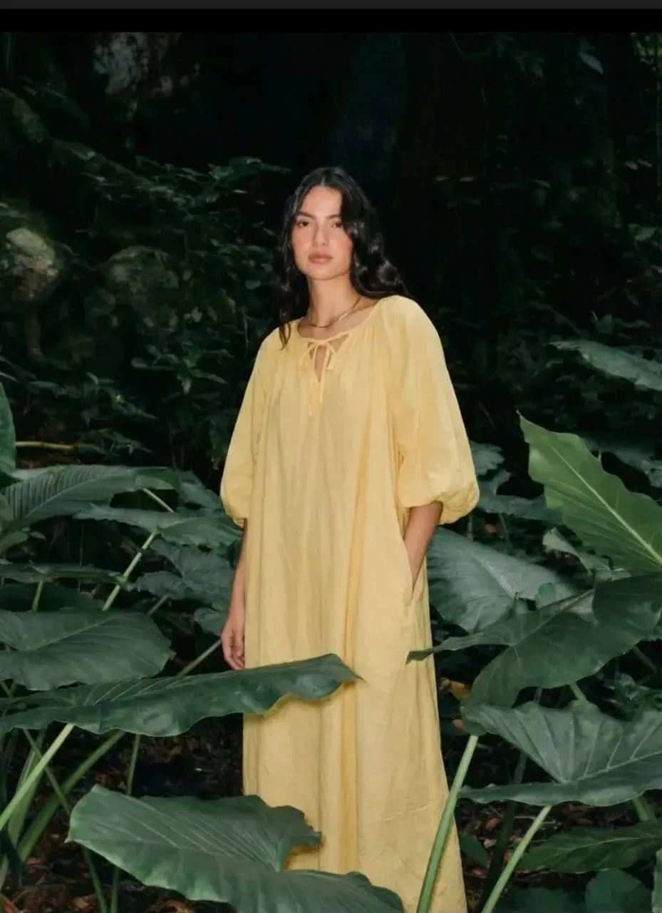 Une personne en robe jaune se tient au milieu de grandes feuilles tropicales vertes.