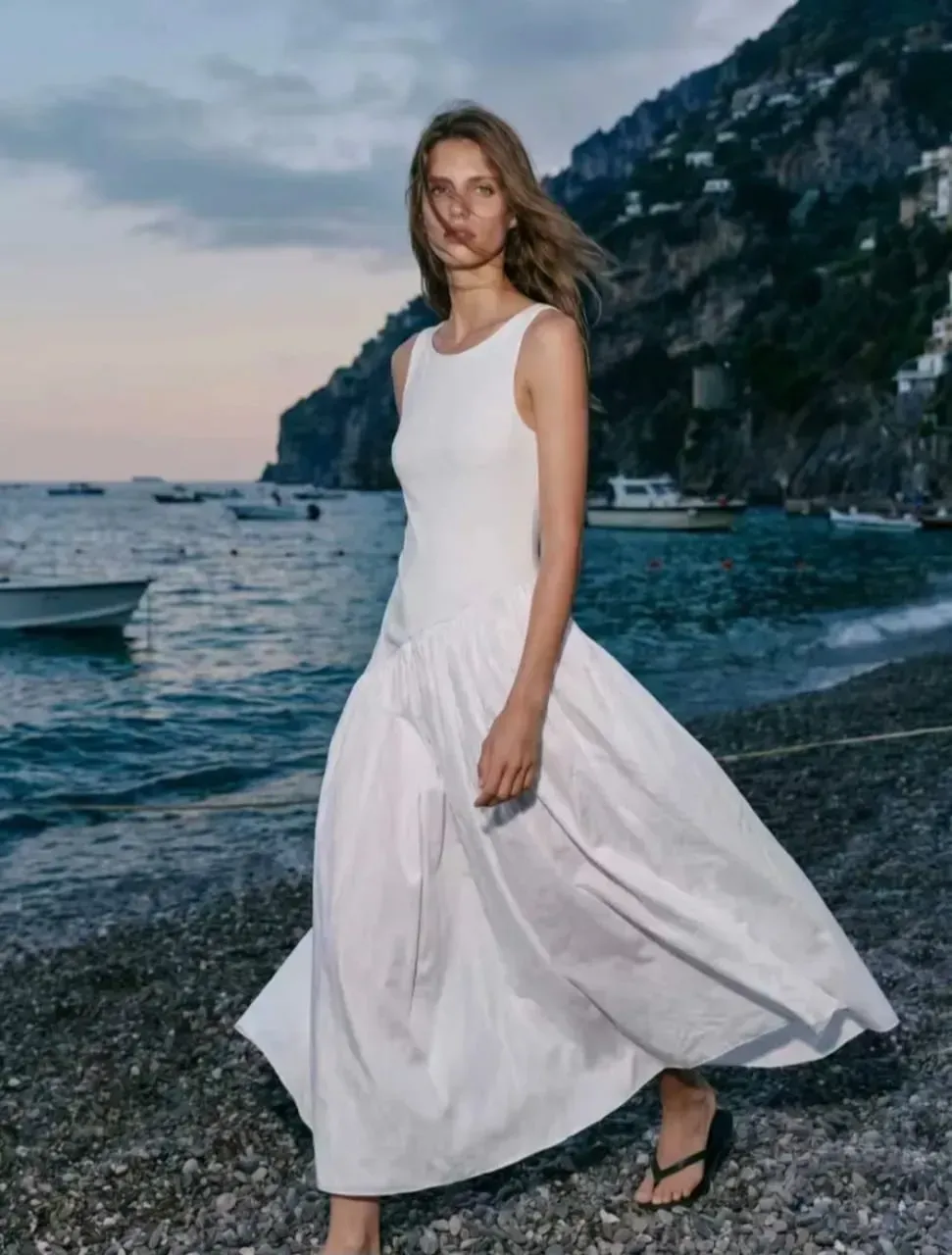 Une femme en robe blanche fluide se tient debout sur une plage rocheuse au coucher du soleil.