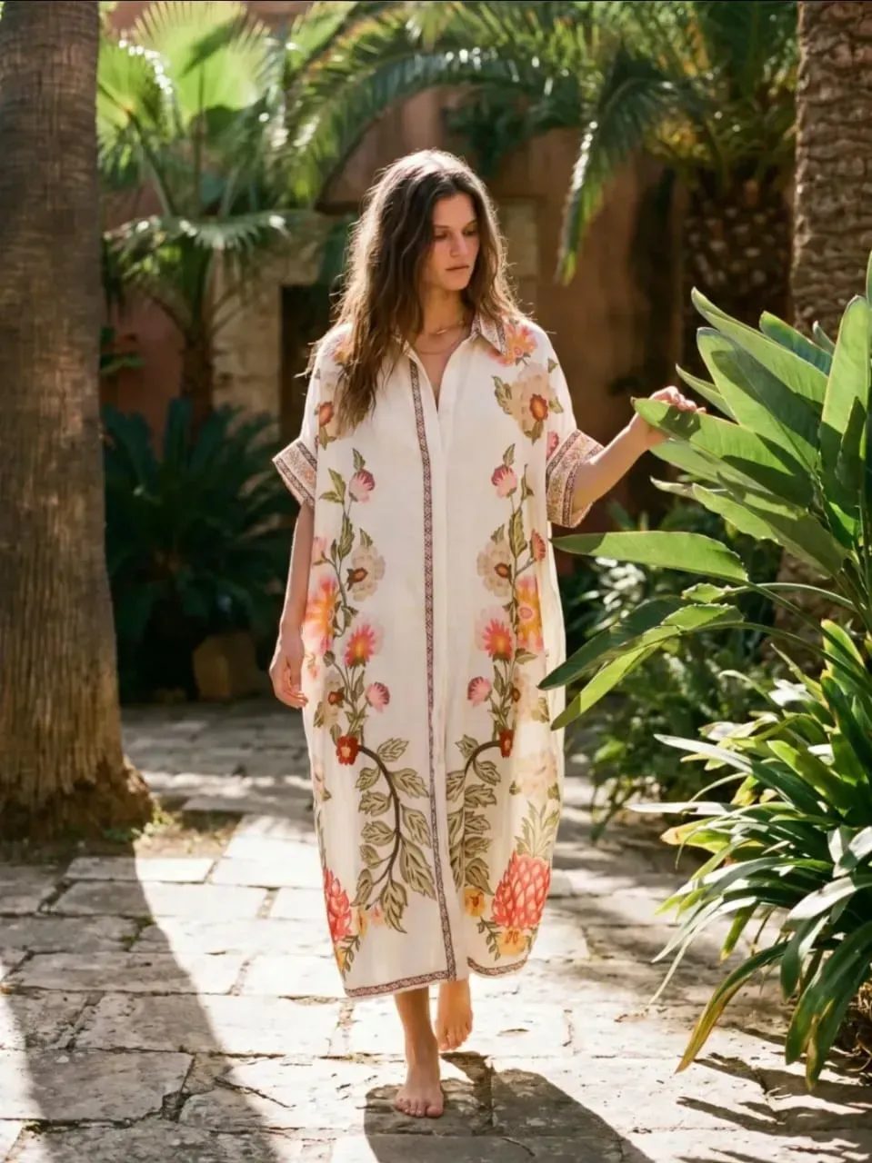 Une femme en kimono blanc à fleurs marche pieds nus sur un chemin de jardin ensoleillé bordé de palmiers.
