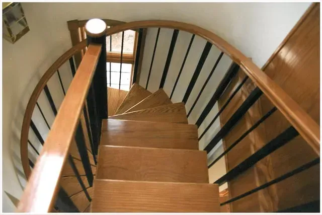 Curved wooden staircase viewed from above, with dark railings and a small window at the landing