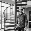 Worker in hard hat standing beside a spiral staircase inside a modern building.