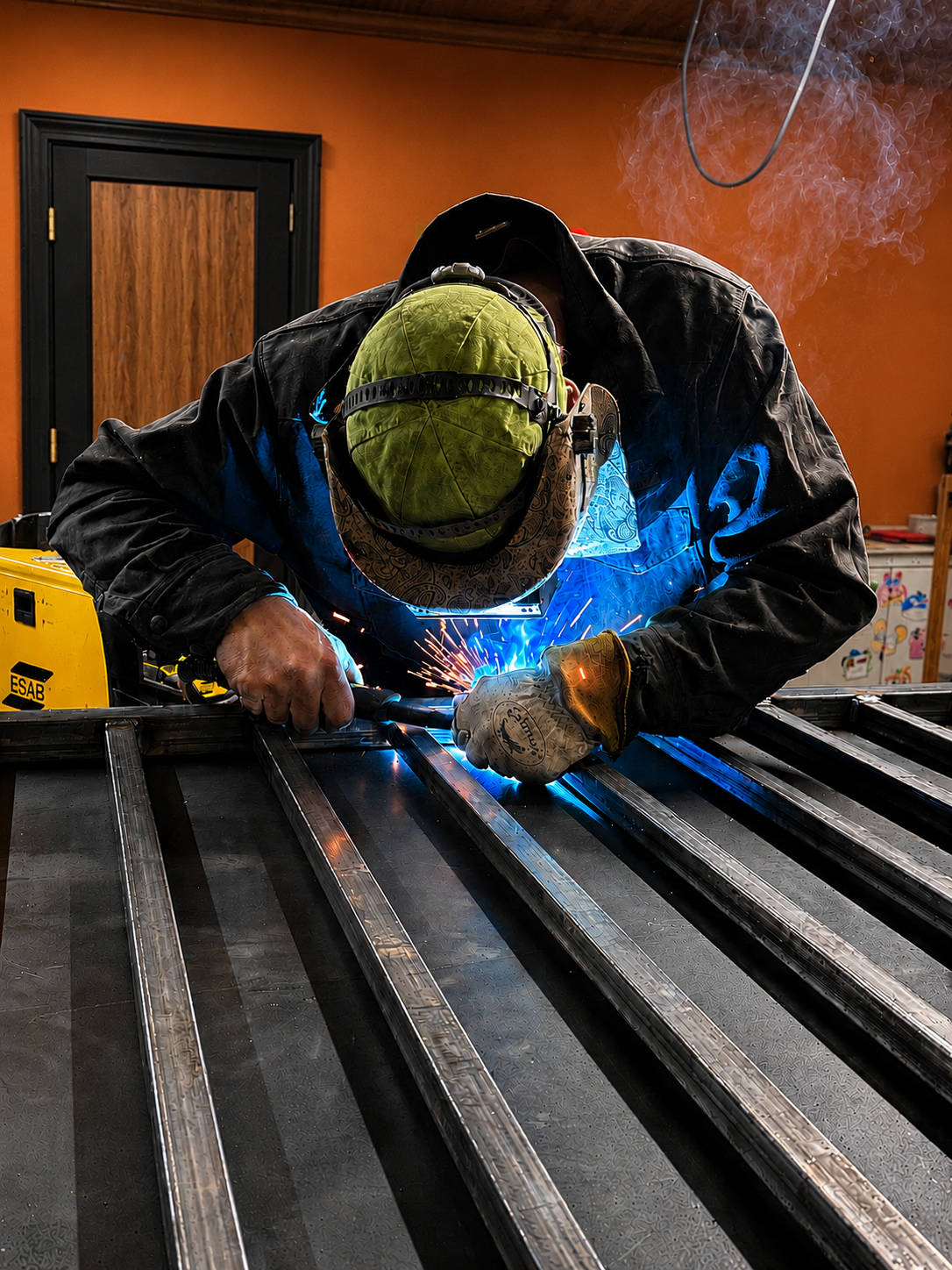 Welder in a green hood and blue jacket welding metal beams, sparks flying in a workshop.