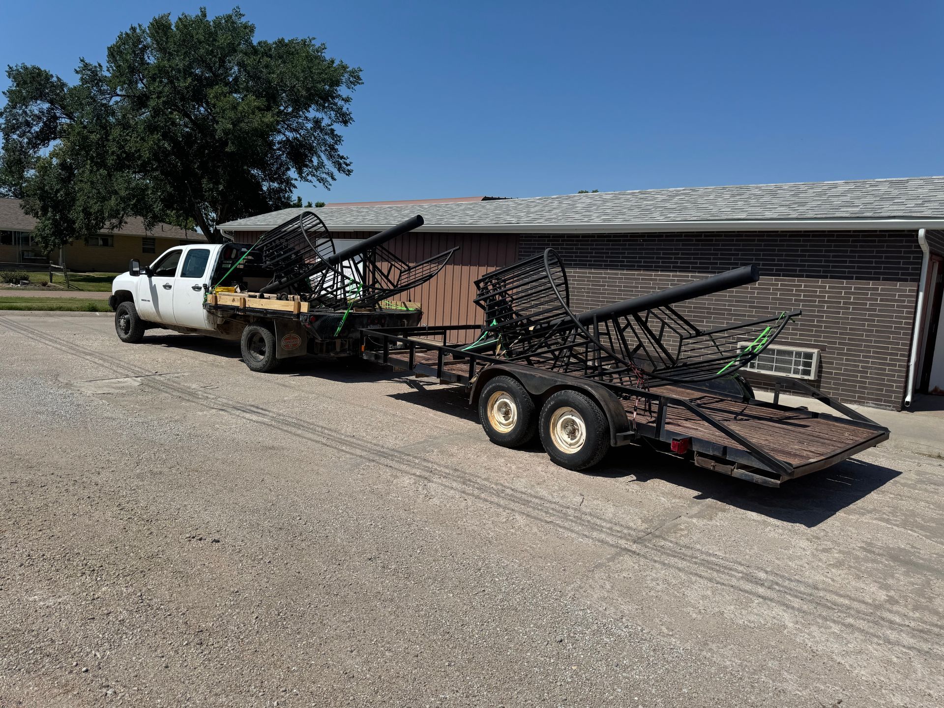 White pickup truck towing two flatbed trailers loaded with folded green metal structures beside a brick building.