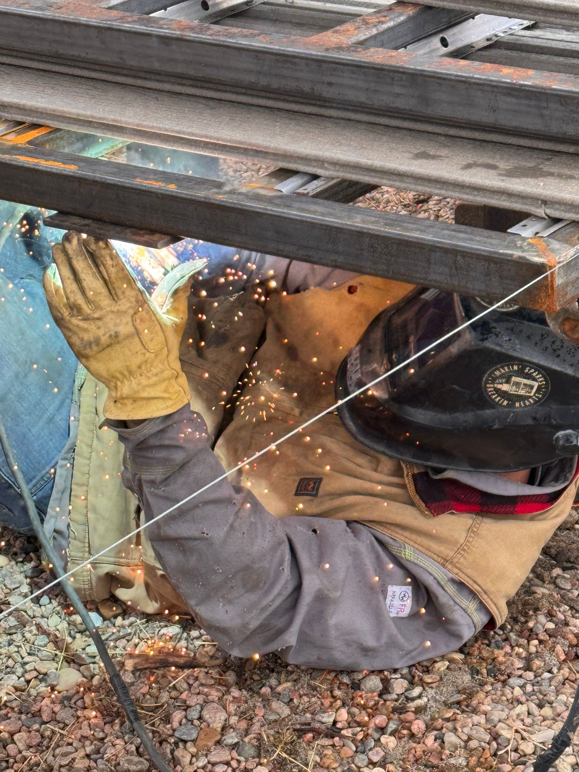 Worker in gloves and hard hat crouches under metal beams, using an angle grinder with sparks flying.