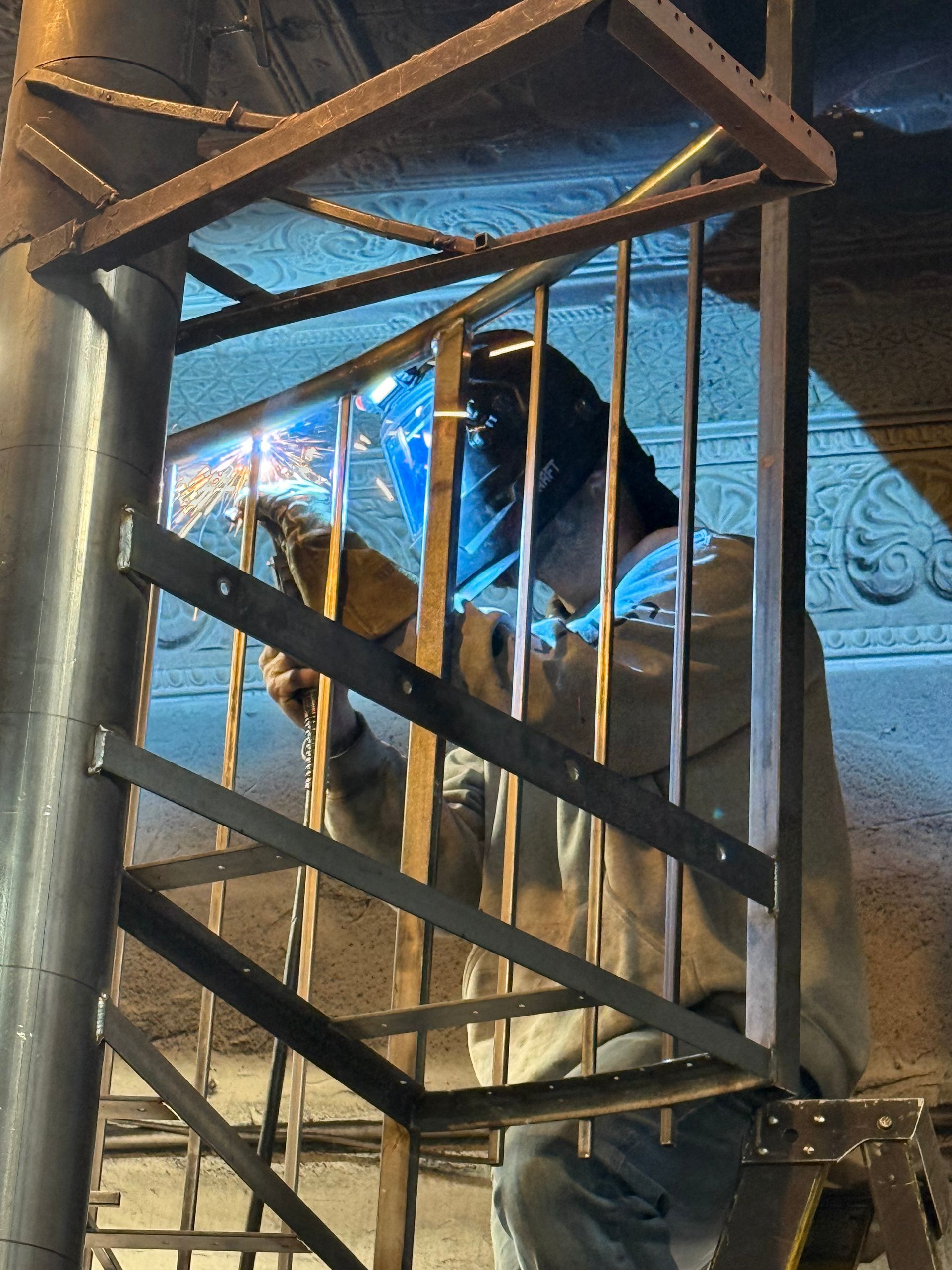 Person in a black jacket and face shield standing inside a wooden crate under a metal frame.