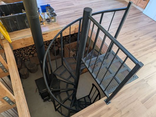 Black metal spiral staircase descending through a wooden deck opening with a handrail and mesh steps