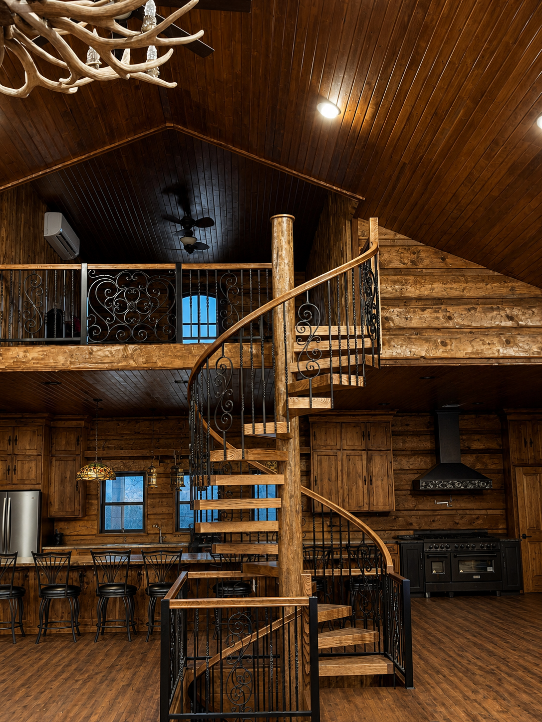 Rustic wooden lodge interior with a spiral staircase, loft balcony, and mounted antlers.