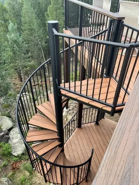 Spiral wooden stairway with black metal railing on a cliffside deck overlooking a forest.