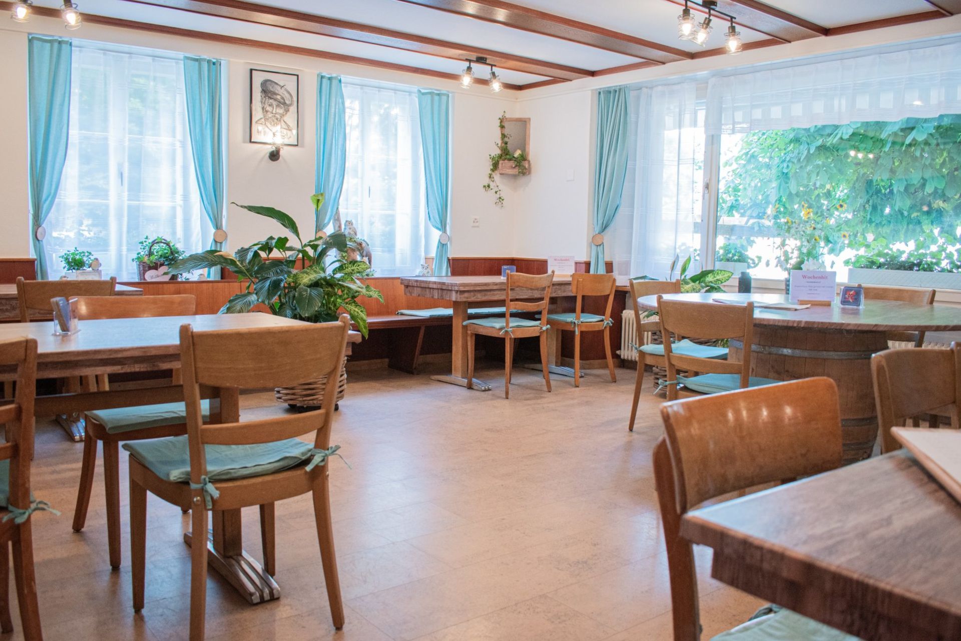 Heller Gastraum eines Cafés mit Holztischen, Stühlen, blauen Vorhängen und großen, sonnendurchfluteten Fenstern