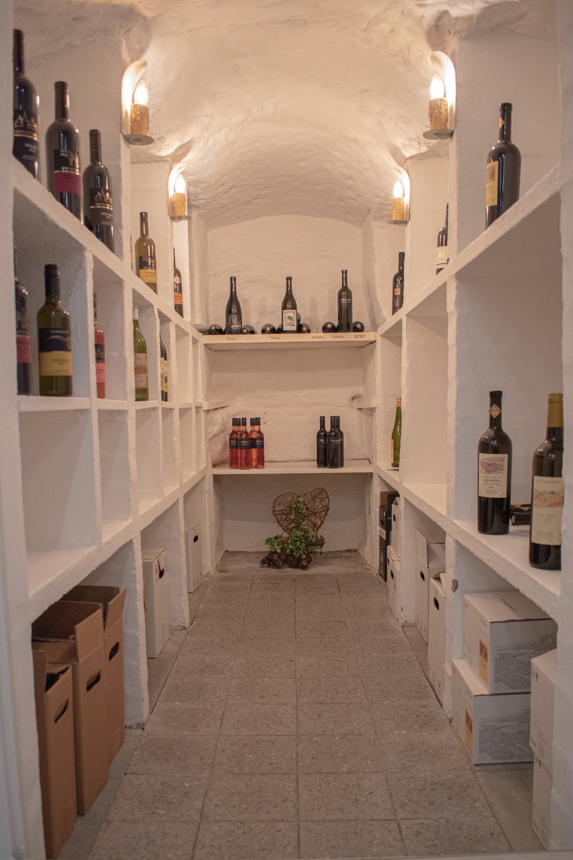 Schmaler Weinkeller mit Flaschen auf weißen Regalen und gewölbter Decke mit Beleuchtung