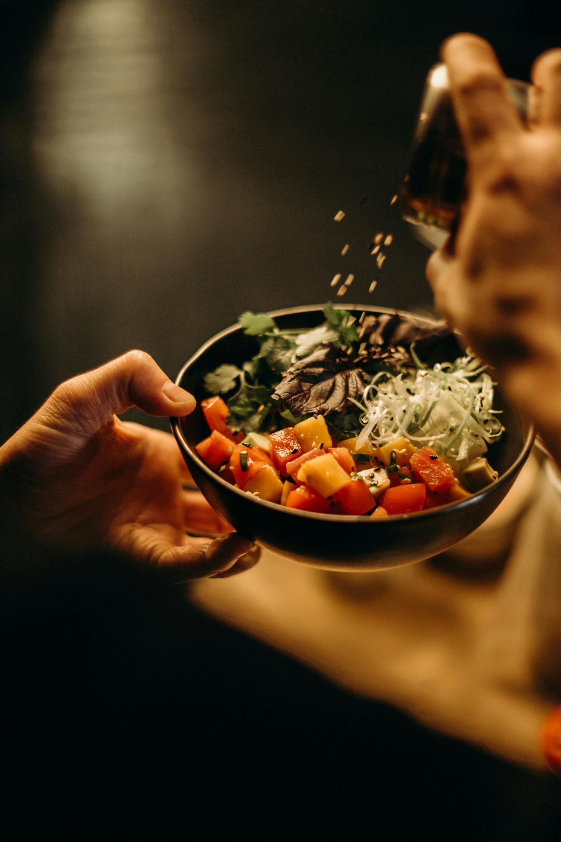 A hand holds a bowl of chopped fruit and greens as someone sprinkles seeds over it from a small container.