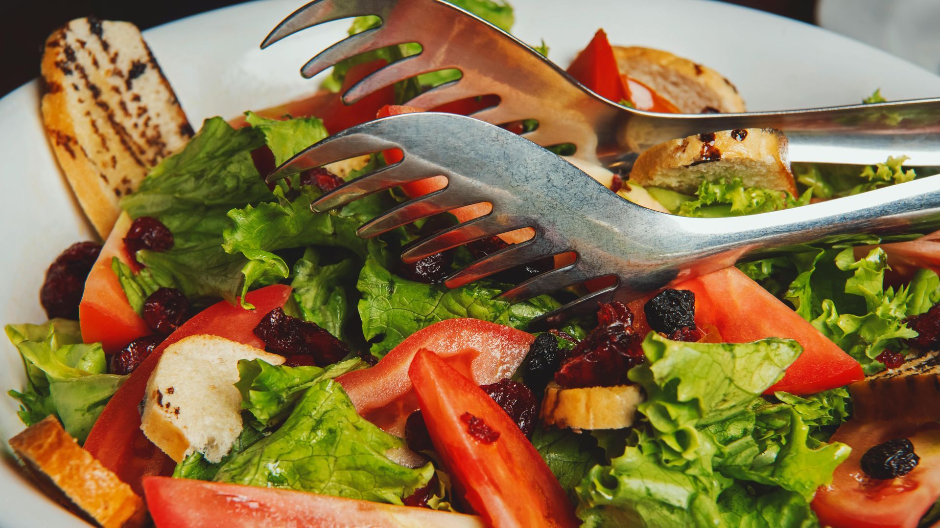 Salad with lettuce, tomatoes, dried cranberries, bread, and serving tongs.