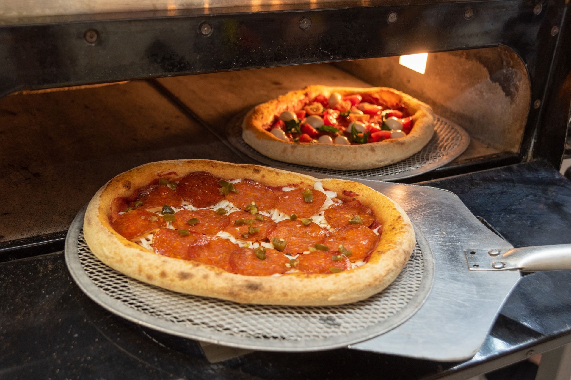 Two pizzas baking in an oven, one with pepperoni, the other with vegetables, on metal pizza screens.