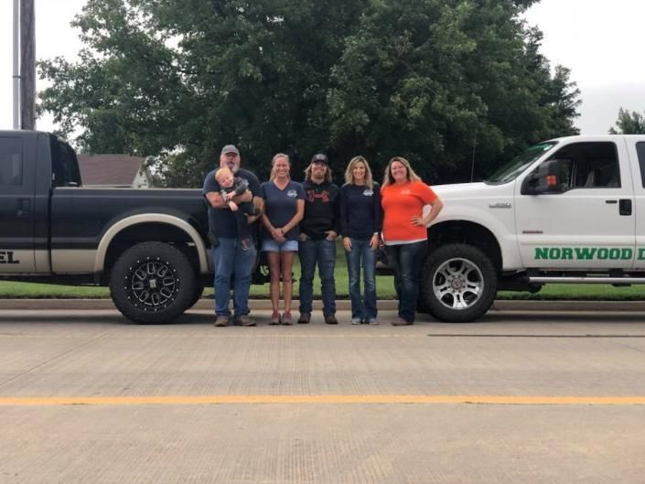 A group of five people stand side-by-side between a black pickup truck and a white work truck parked on a paved road.