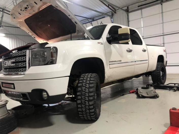 A white GMC Sierra pickup truck with its hood open, lifted on a vehicle hoist inside a workshop.