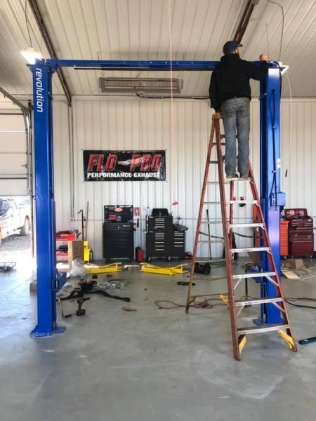 A person on a ladder installs an overhead support beam on a blue two-post automotive lift inside a garage.