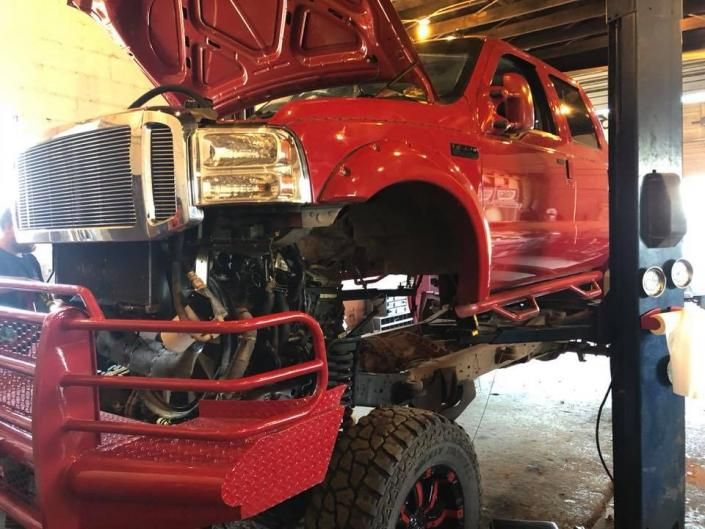 A bright red lifted pickup truck with its hood open, positioned on a vehicle service lift inside a repair shop.
