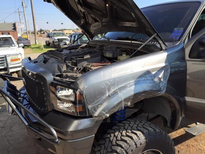 A dark gray lifted truck with its hood open, parked on a paved lot with protective plastic covering the front fender.