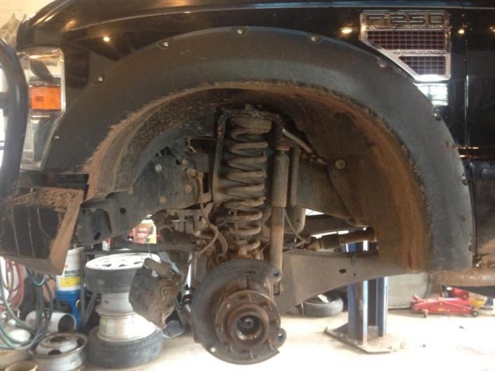 A close-up of a vehicle's front suspension, showing a coil spring, shock absorber, and wheel hub in an auto shop setting.